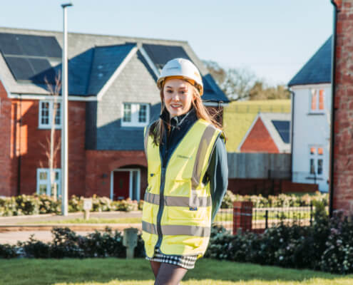 Photo of young woman standing in front of new houses wearing a hard hat and high viz jacket.