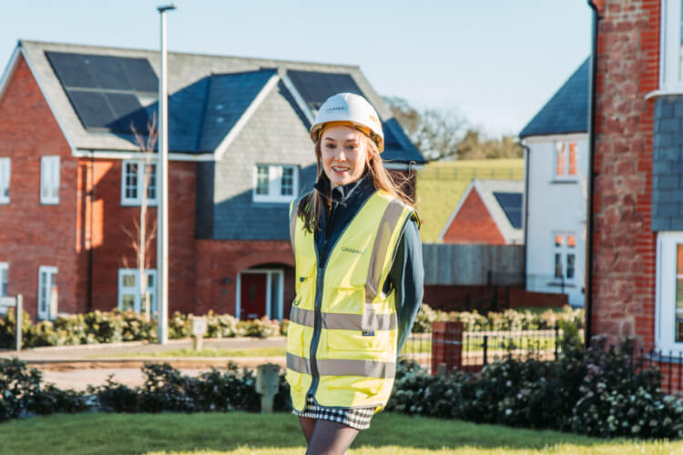 Photo of young woman standing in front of new houses wearing a hard hat and high viz jacket.